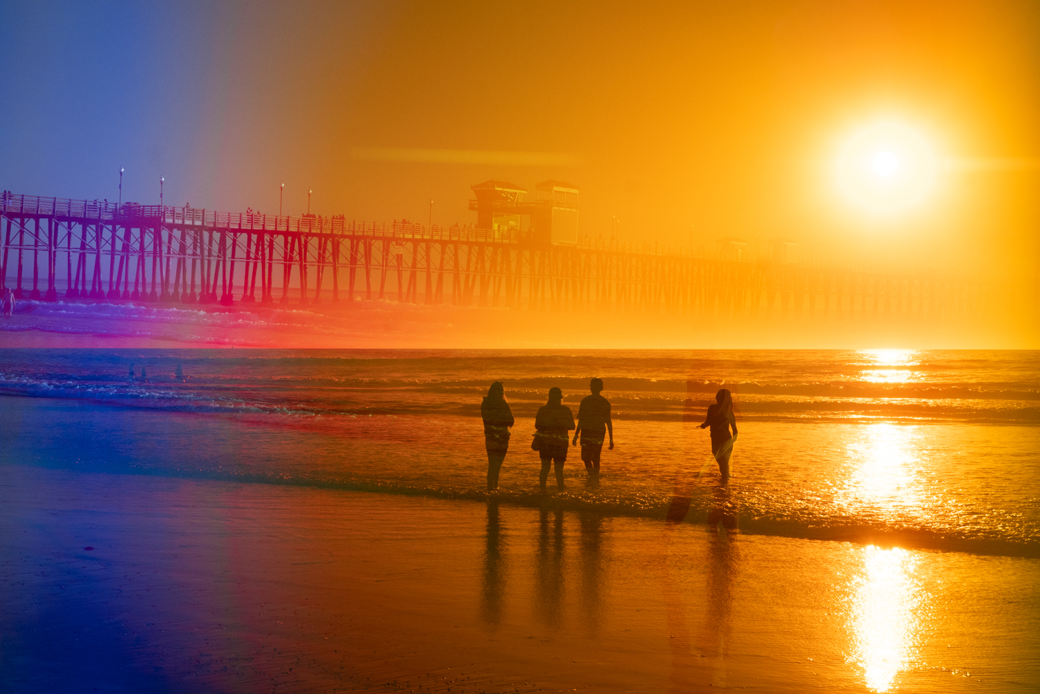 Prism photo look at both Oceanside pier on left side and 4 people standing in the ocean at sunset.
